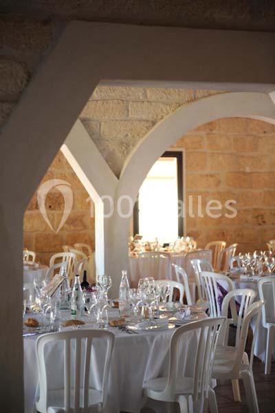 Salle de réception avec tables rondes dressées, nappes blanches, verres et chaises en plastique, murs en pierre apparente.