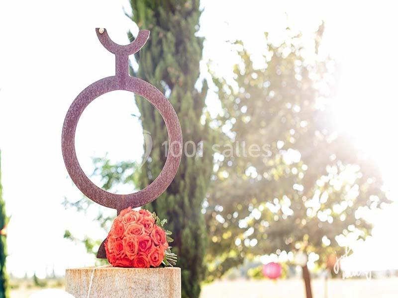 Sculpture métallique circulaire sur un socle, accompagnée d'un bouquet de roses rouges, en extérieur ensoleillé.
