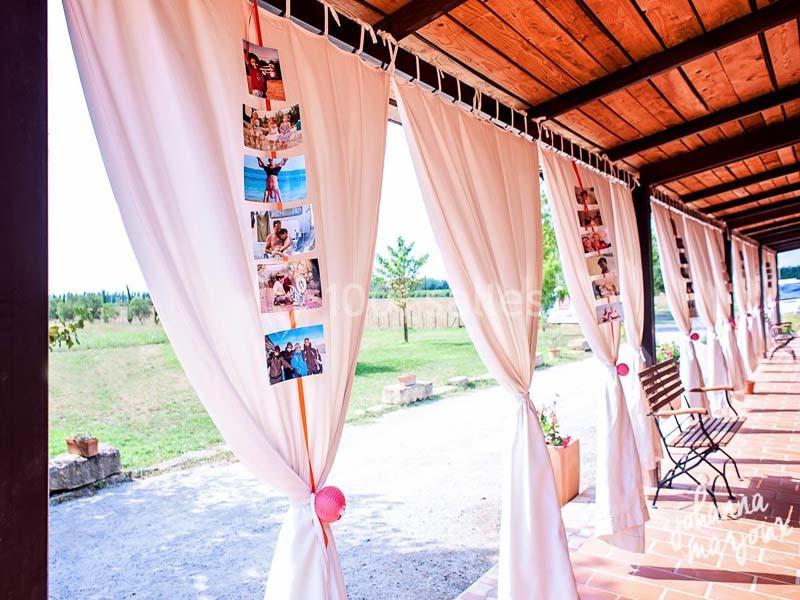 Rideaux blancs décorés de photos suspendus sous une pergola en bois, avec des chaises et un paysage champêtre en arrière…