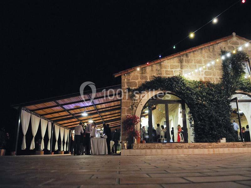 Façade d'un bâtiment en pierre éclairé la nuit, avec des invités rassemblés sous une terrasse couverte.