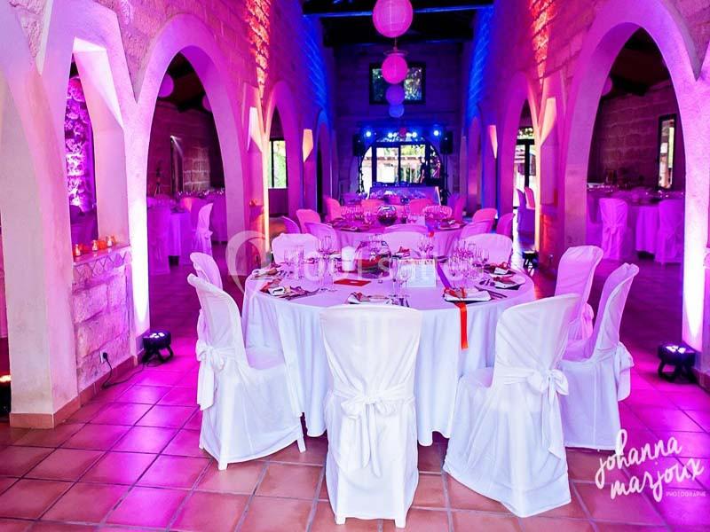 Salle de réception décorée avec des tables rondes habillées de nappes blanches et éclairage d'ambiance coloré.