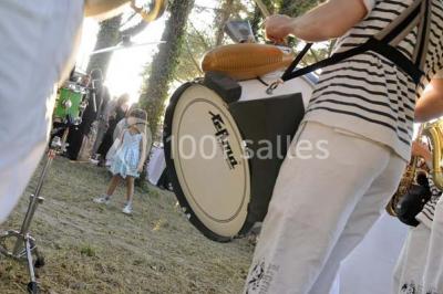 Des musiciens en tenue blanche et rayée jouent des percussions en extérieur, près d'une petite fille.