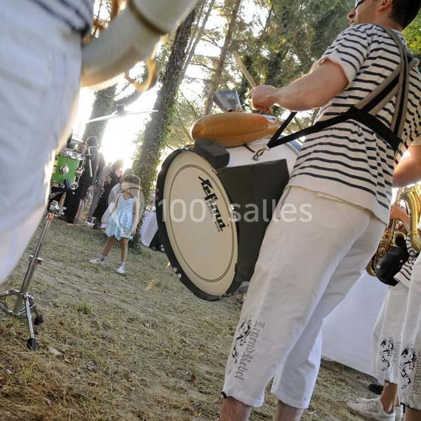 Des musiciens en tenue blanche et rayée jouent des percussions en extérieur, près d'une petite fille.