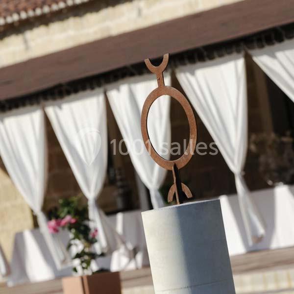 Sculpture en métal sur un socle devant un bâtiment avec des rideaux blancs et des plantes en arrière-plan.