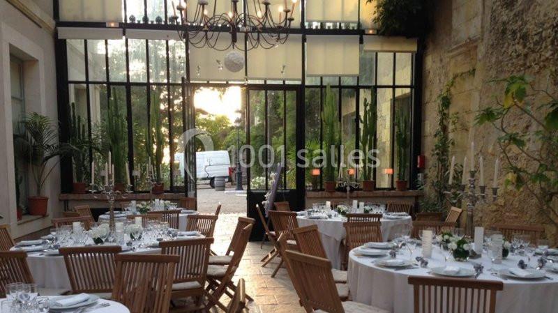 Salle lumineuse avec grandes baies vitrées, tables rondes dressées et chaises en bois, entourées de plantes vertes.