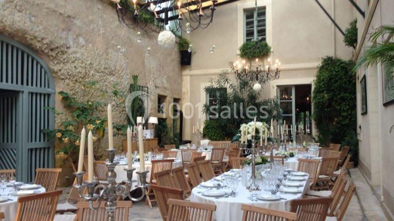 Salle lumineuse avec tables dressées, chaises en bois, chandeliers et plantes, sous une verrière élégante.