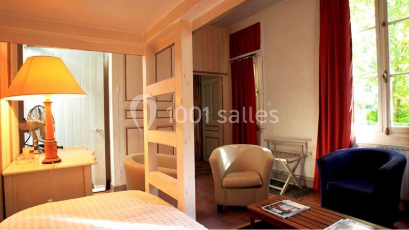 Chambre lumineuse avec lit, fauteuils, table basse, lampadaire et rideaux rouges, vue sur un jardin à travers la fenêtre.