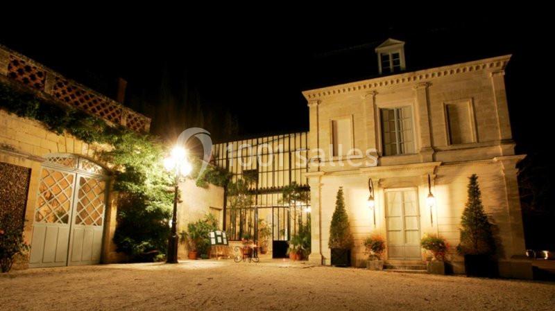 Cour d'une maison en pierre éclairée la nuit, avec des plantes, des portes vitrées et des lampadaires allumés.