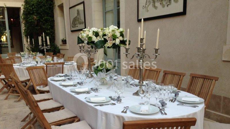 Table élégamment dressée avec nappes blanches, chandeliers, fleurs et vaisselle, dans une cour intérieure lumineuse.