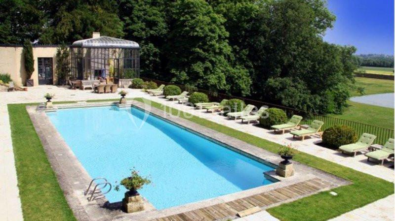Piscine extérieure entourée de chaises longues, végétation et terrasse, avec vue sur un paysage champêtre.