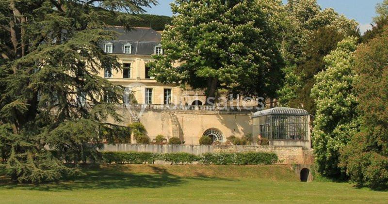 Façade d'un bâtiment ancien entouré d'arbres, avec une serre vitrée et une pelouse au premier plan.
