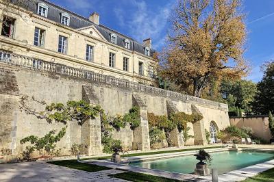 Façade d'un bâtiment ancien en pierre avec une piscine et un jardin arboré sous un ciel bleu.