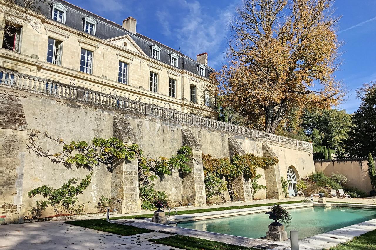 Façade d'un bâtiment ancien en pierre avec une piscine et un jardin arboré sous un ciel bleu.