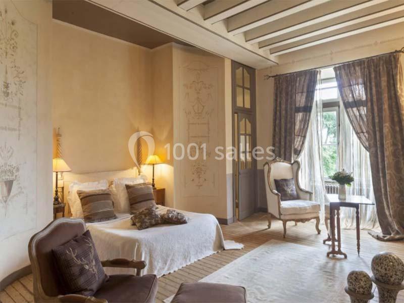 Chambre lumineuse avec lit double, fauteuils, rideaux élégants et vue sur un jardin à travers une grande fenêtre.