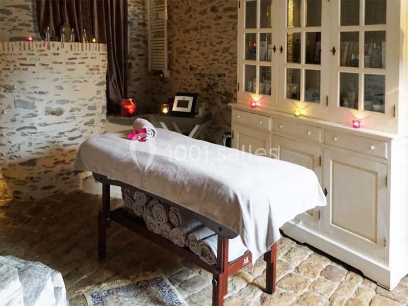 Table de massage recouverte d'un drap blanc dans une pièce en pierre avec bougies allumées et mobilier rustique.