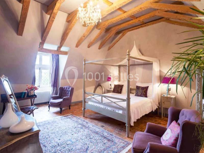 Chambre lumineuse avec lit à baldaquin, poutres apparentes, fauteuils violets, tapis et fenêtre donnant sur l'extérieur.