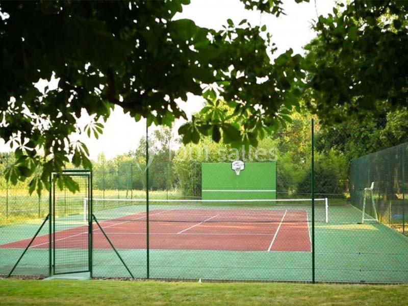 Terrain de tennis extérieur entouré de verdure, avec un mur d'entraînement vert au fond.