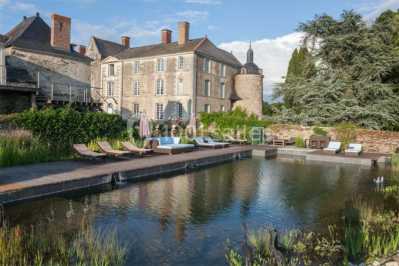 Bassin naturel entouré de végétation avec des transats en bois, devant un bâtiment ancien en pierre sous un ciel dégagé.