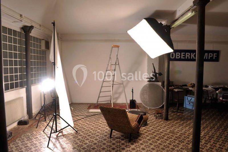 Studio photo avec éclairages, échelle, chaise en rotin et accessoires sur un sol à motifs géométriques.