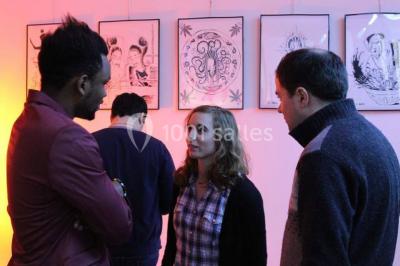 Exposition artistique dans une salle avec des œuvres colorées accrochées aux murs et des photos disposées sur une table.