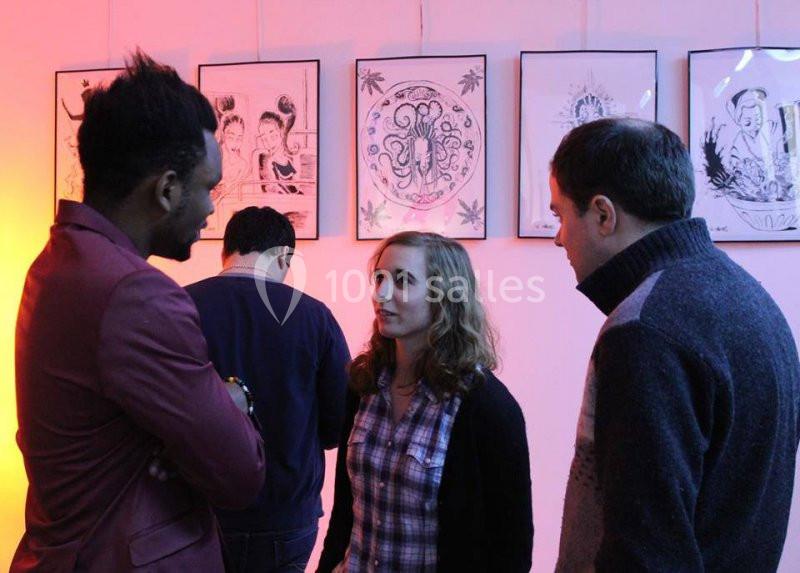 Trois personnes discutent dans une galerie d'art avec des illustrations encadrées accrochées au mur en arrière-plan.