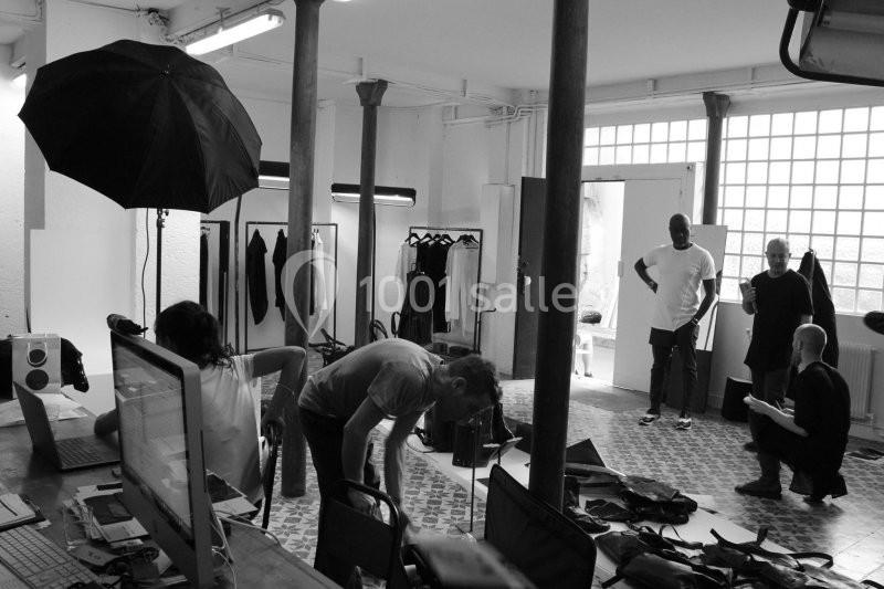 Photographes et mannequins travaillent dans un studio lumineux avec équipements et vêtements en arrière-plan.