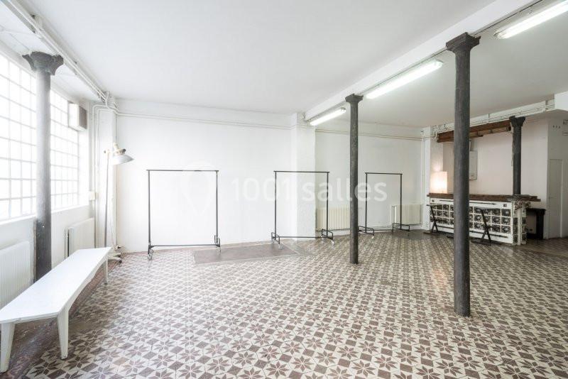 Salle lumineuse avec sol en carreaux de ciment, murs blancs, grandes fenêtres, banc, portants et piliers métalliques.