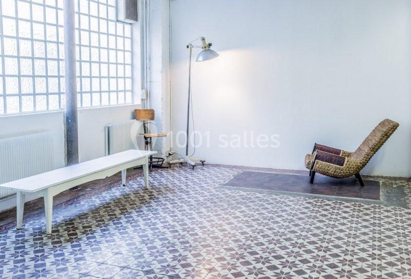 Salle lumineuse avec sol en carreaux de ciment, chaise en rotin, banc blanc et lampe sur pied près d'un mur blanc.