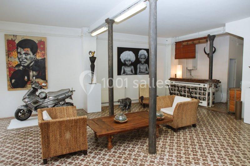 Salon avec fauteuils en rotin, table basse en bois, œuvres d'art au mur, scooter exposé et sol en carreaux décoratifs.