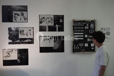 Exposition artistique dans une salle avec des œuvres colorées accrochées aux murs et des photos disposées sur une table.