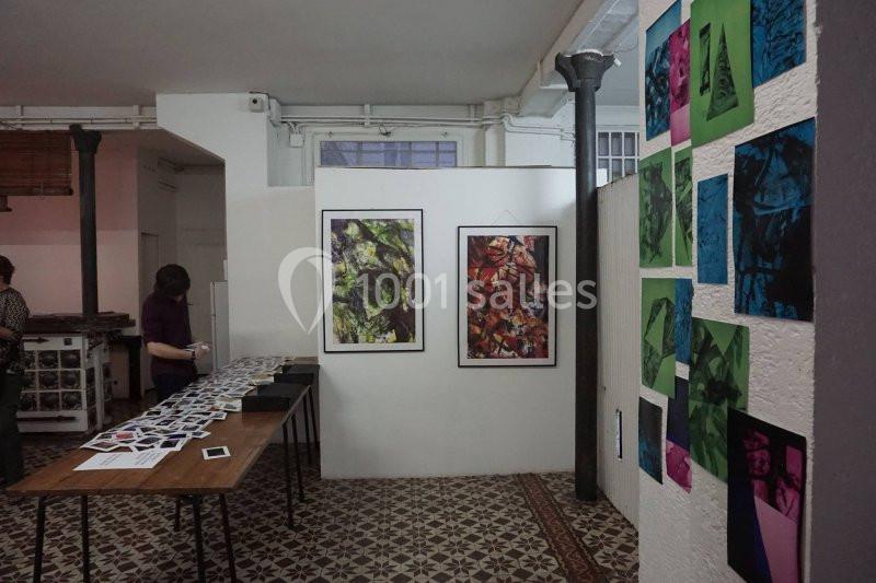 Exposition artistique dans une salle avec des œuvres colorées accrochées aux murs et des photos disposées sur une table.