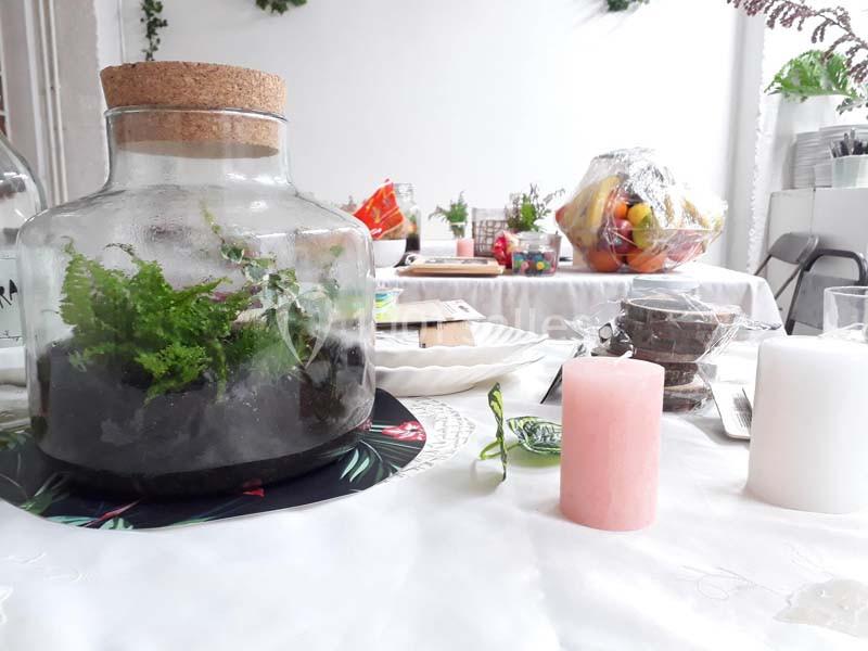 Terrarium en verre avec des fougères sur une table décorée, entouré de bougies et d'objets divers.