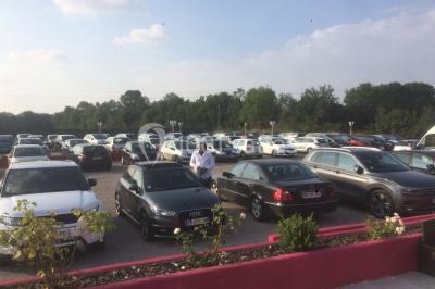 Parking extérieur rempli de voitures, avec un homme debout près d'une Audi noire sous un ciel dégagé.