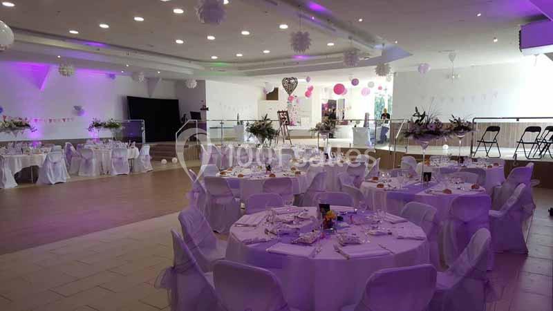 Salle de réception décorée avec des tables rondes dressées, des chaises blanches et un éclairage violet.