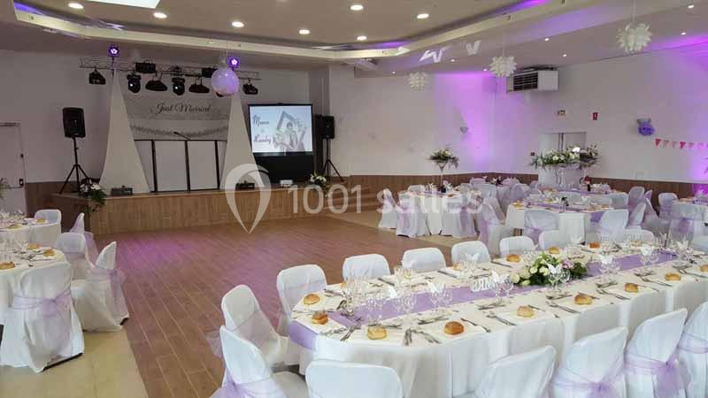 Salle de réception décorée pour un événement, avec tables dressées, nappes blanches et éclairage violet.