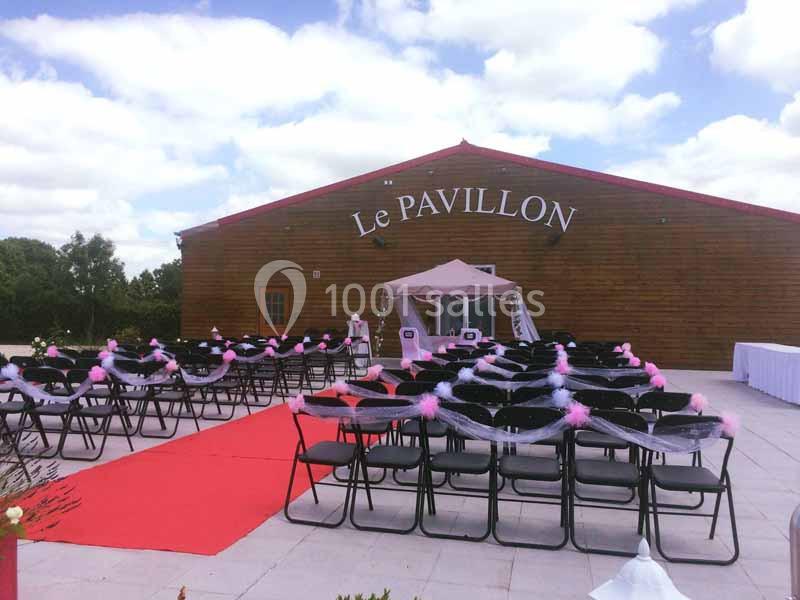 Chaises alignées avec décorations roses et blanches devant un bâtiment en bois marqué ’Le Pavillon’, sous un ciel…