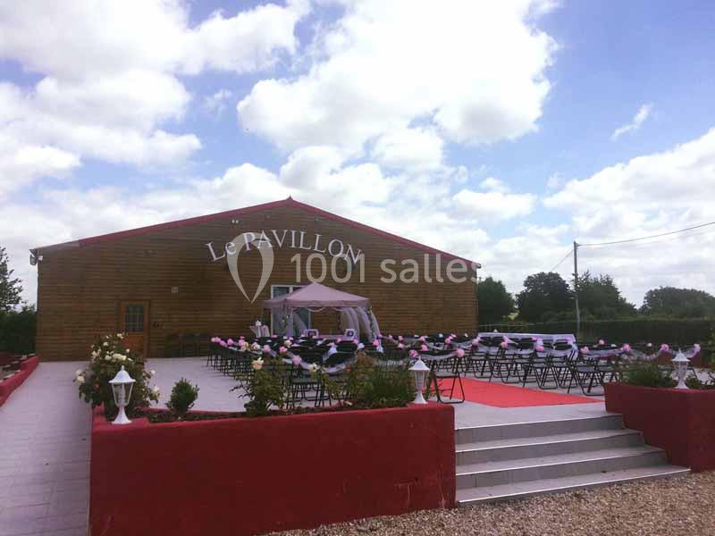 Salle de réception en bois avec terrasse aménagée, chaises décorées et tapis rouge, sous un ciel partiellement nuageux.