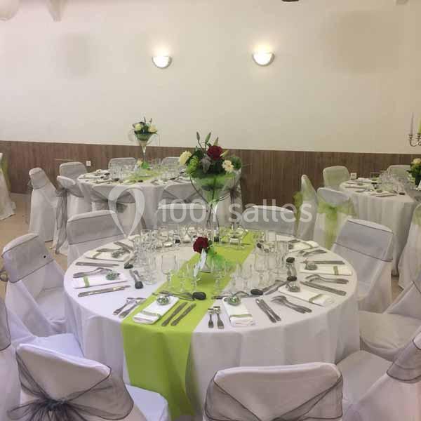 Salle décorée pour un événement avec des tables rondes dressées, nappes blanches, chemins de table verts et chaises houssées.