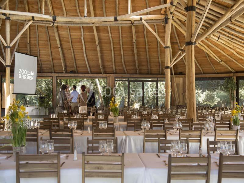 Salle de réception en bois avec tables dressées, chaises en bois et un écran de projection allumé au fond.