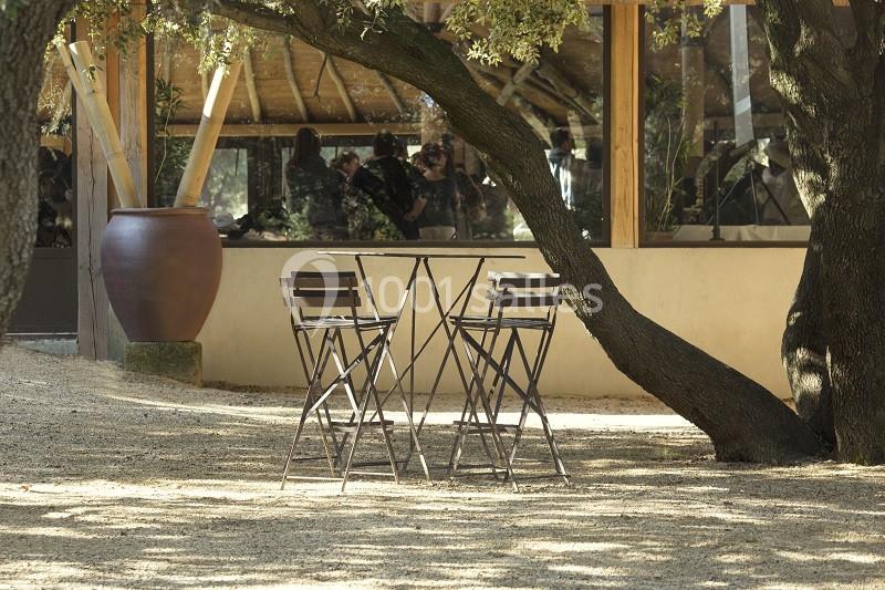 Table et chaises en métal noir installées sous des arbres, près d'une grande fenêtre reflétant l'extérieur.