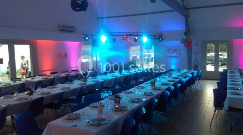 Salle décorée pour un événement, avec des tables dressées, des chaises bleues et un éclairage coloré.