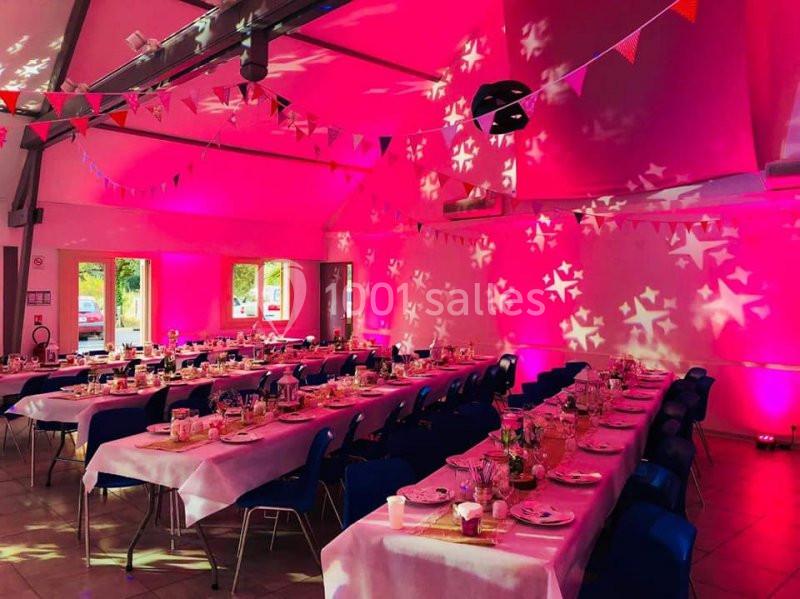 Salle décorée pour un événement festif, avec tables dressées, guirlandes colorées et éclairage rose.
