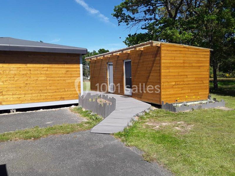 Deux bâtiments en bois reliés par une passerelle, entourés de pelouse et d'arbres sous un ciel bleu.