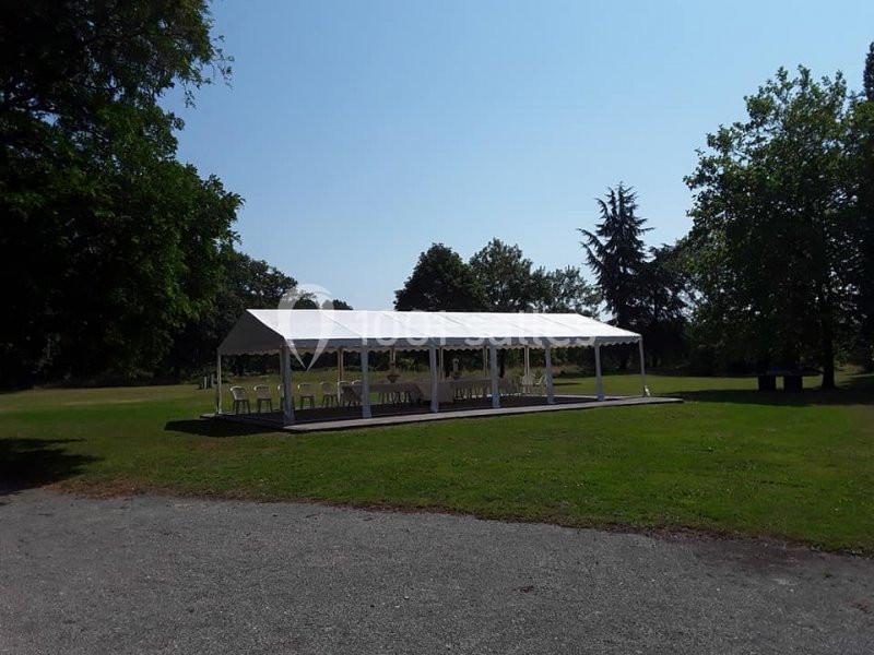 Tente blanche montée dans un espace vert, entourée d'arbres, avec des chaises disposées à l'intérieur.
