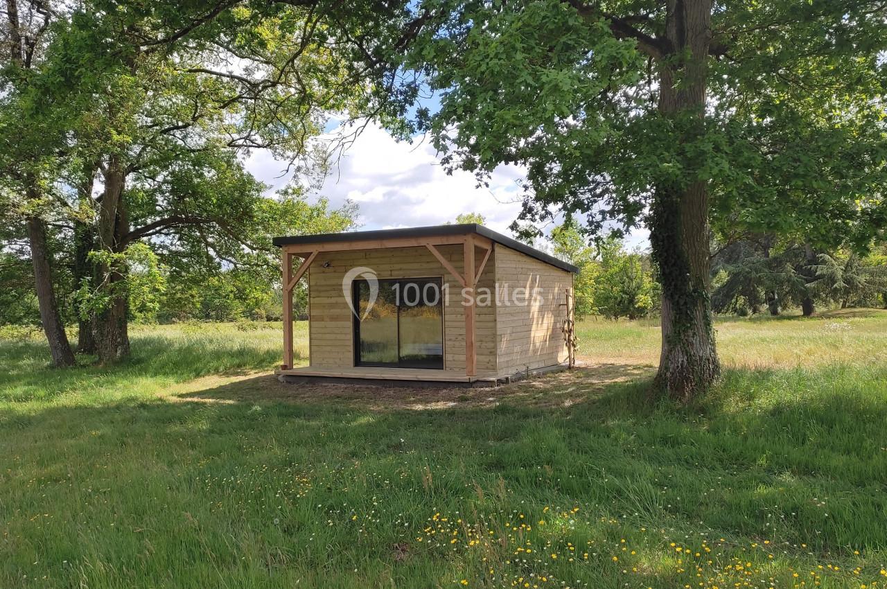 Petite maison en bois avec une grande baie vitrée, située dans un champ verdoyant entouré d'arbres.