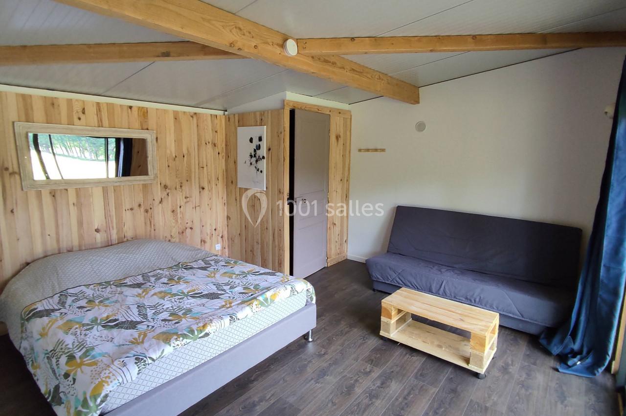 Chambre lumineuse avec lit double, canapé, table basse en bois, murs en bois clair et poutres apparentes.