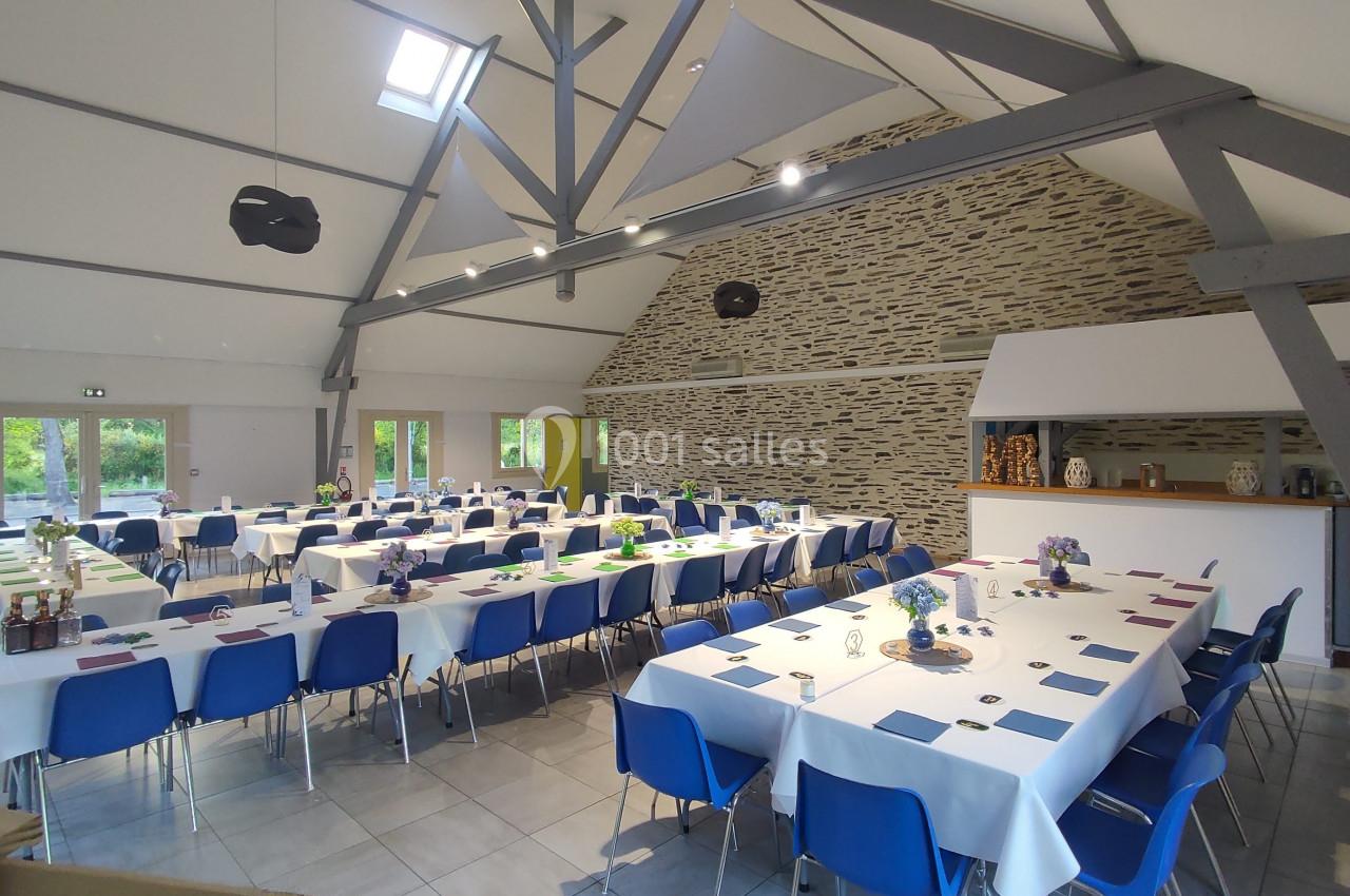 Salle lumineuse avec tables dressées en U, nappes blanches, chaises bleues et murs en pierre apparente.