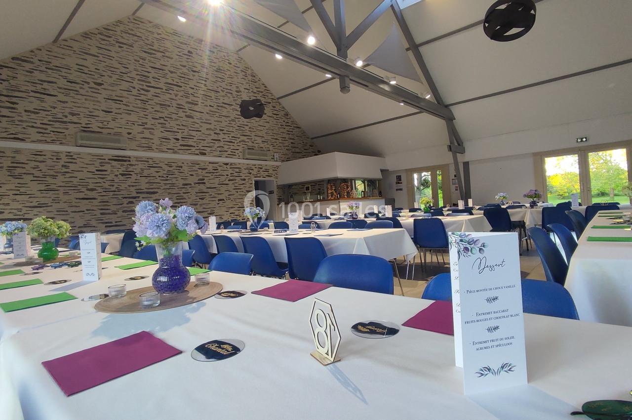 Salle de réception lumineuse avec tables dressées, décorations florales et chaises bleues sous un plafond en poutres…
