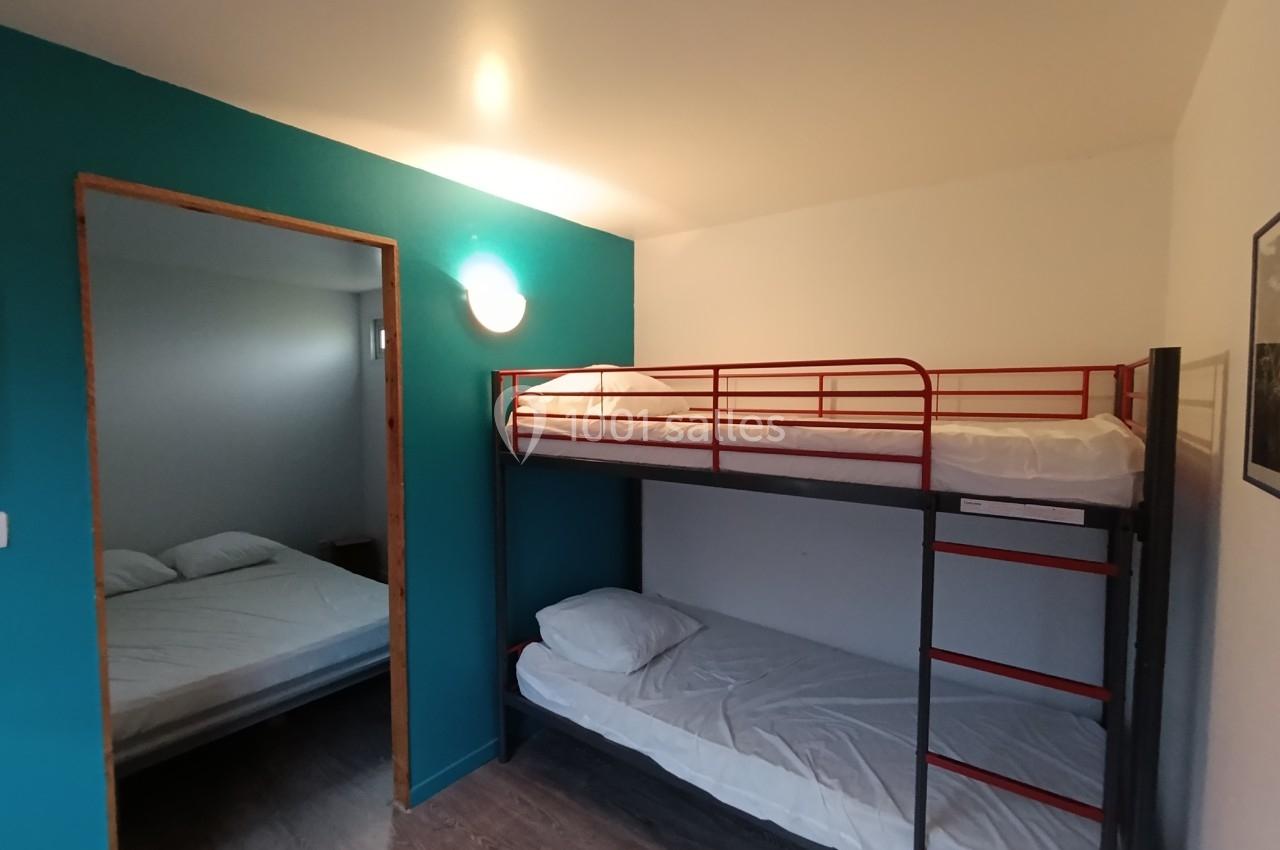 Chambre avec lits superposés en métal rouge, mur bleu, sol en bois et vue sur une autre pièce avec un lit double.