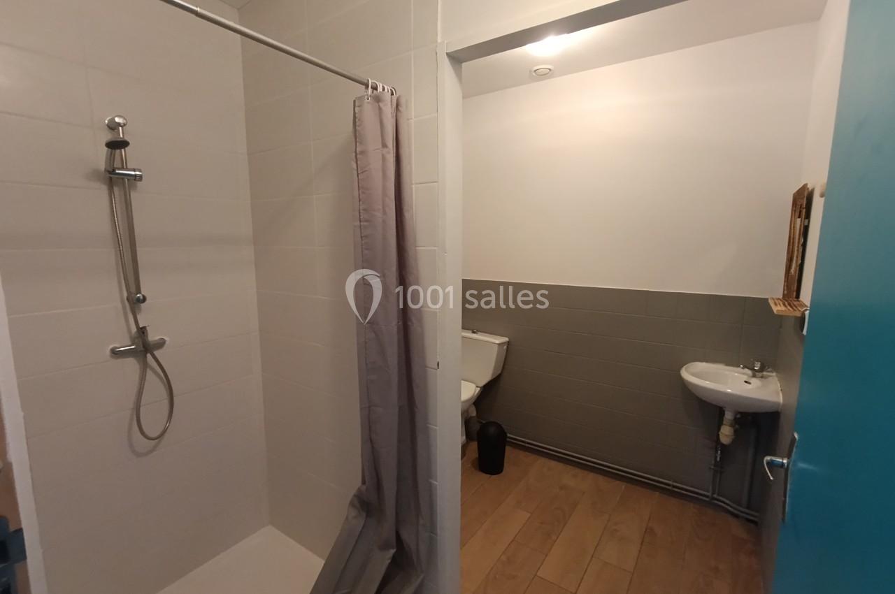 Douche avec rideau gris à gauche, toilettes et lavabo sur mur carrelé gris à droite, sol en parquet.